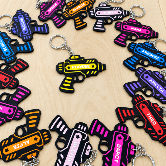 A collection of colorful replica toy gun keychains arranged in a circle on a light wood background. Each keychain is black with bright neon colors (pink, blue, yellow, orange, purple, red) and features a custom name printed on it, such as Darcy, Phoebe, and Theo.